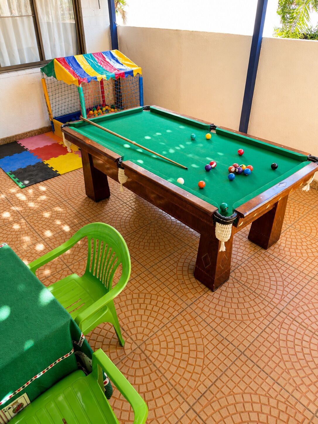 Une table de billard avec des billes, une chaise verte et un sac coloré dans une pièce dont le sol est recouvert de carreaux.