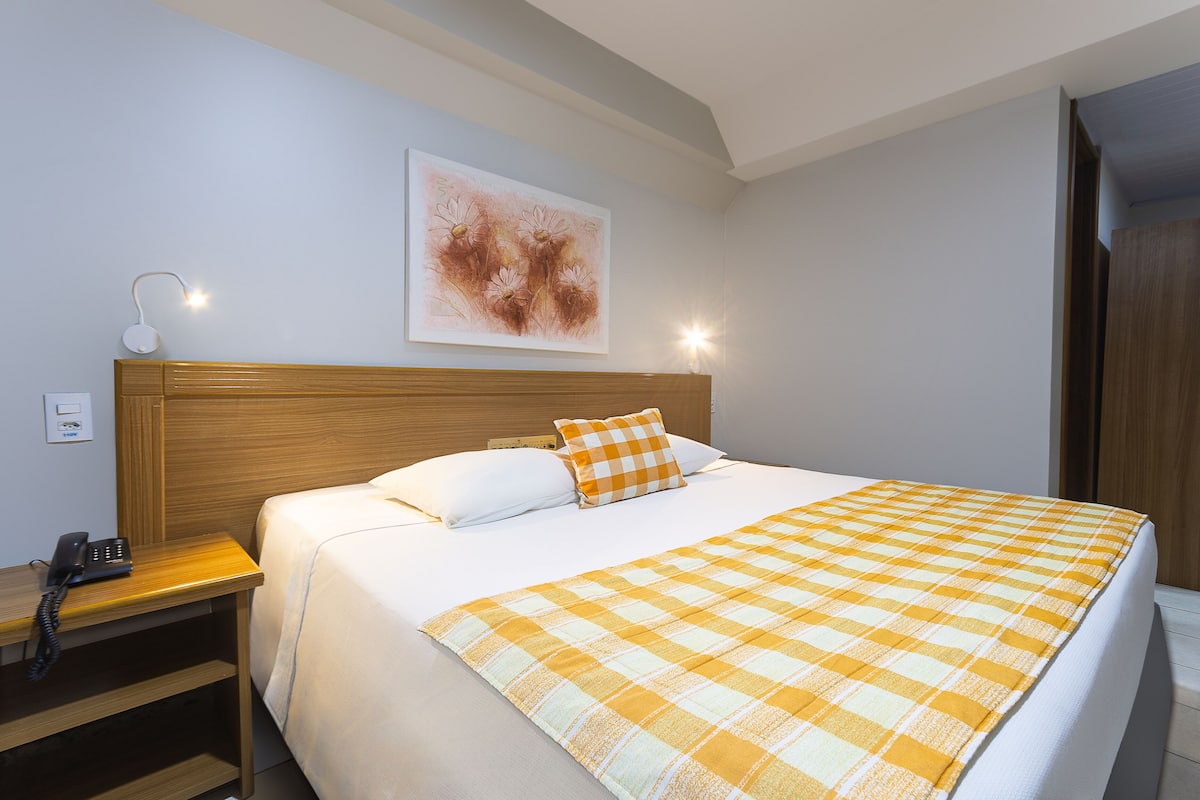 Una cama bien hecha con una colcha a cuadros, un cabecero de madera y una mesita de noche con una lámpara y teléfono.