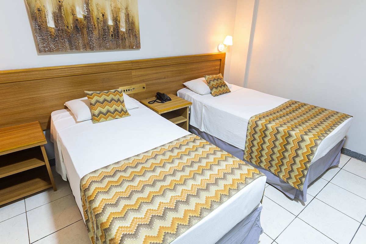Habitación de hotel con dos camas individuales, cada una con colcha y almohadas estampadas. Se observa un cabecero de madera, una mesita de noche con lámpara y un cuadro colgado en la pared.