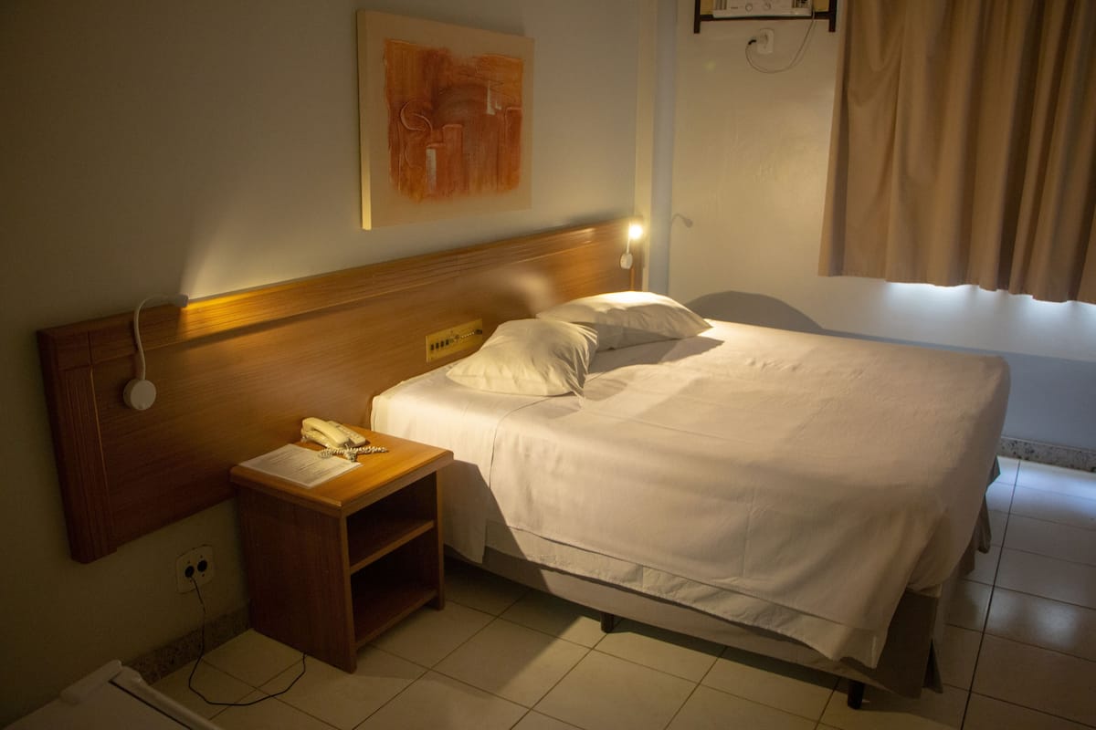 Habitación de hotel con cabecera de madera, mesita de noche con teléfono, lámpara de pared y ventana con cortinas.