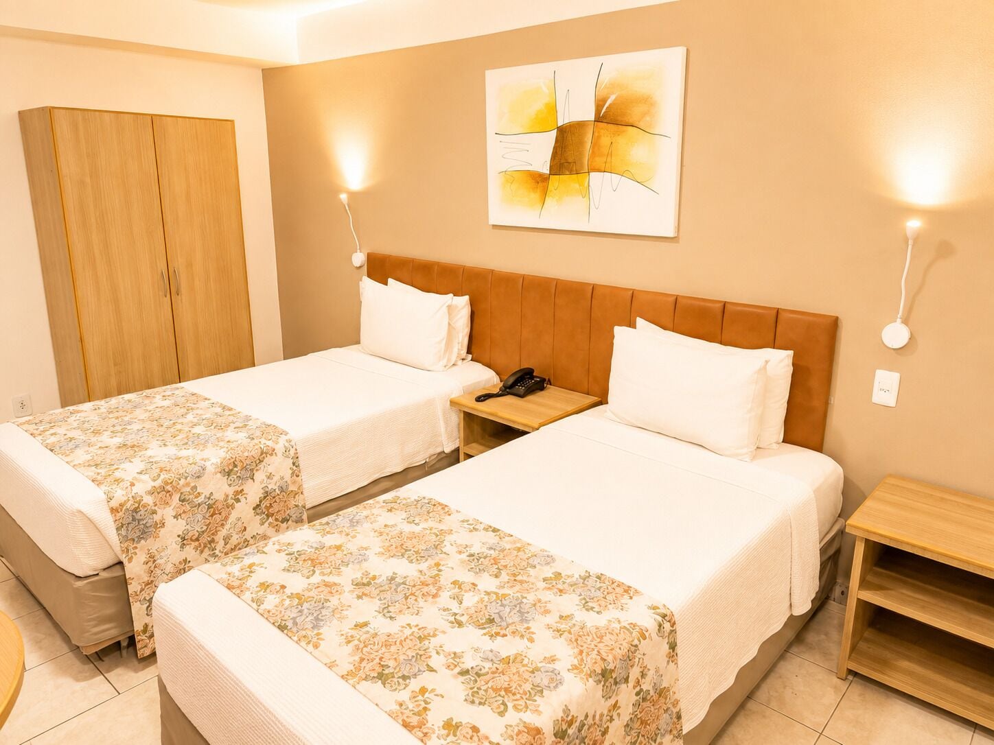 A hotel room with two beds, a wooden wardrobe, a bedside table with a phone, and a wall-mounted abstract painting.