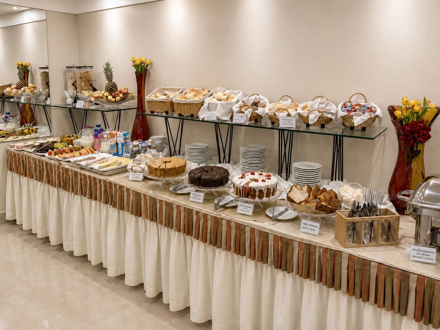 Disposição de buffet com diversos alimentos, incluindo bolos e doces, dispostos em mesas com etiquetas.