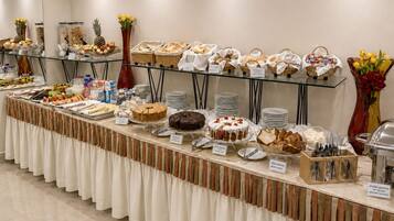 Disposição de buffet com diversos alimentos, incluindo bolos e doces, dispostos em mesas com etiquetas.