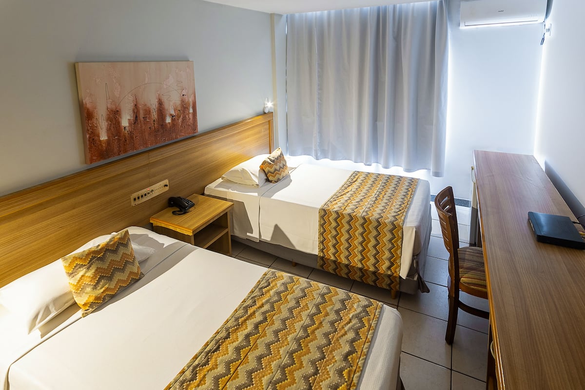 Habitación de hotel con dos camas, cabecera de madera, escritorio, silla y ventana con cortinas.