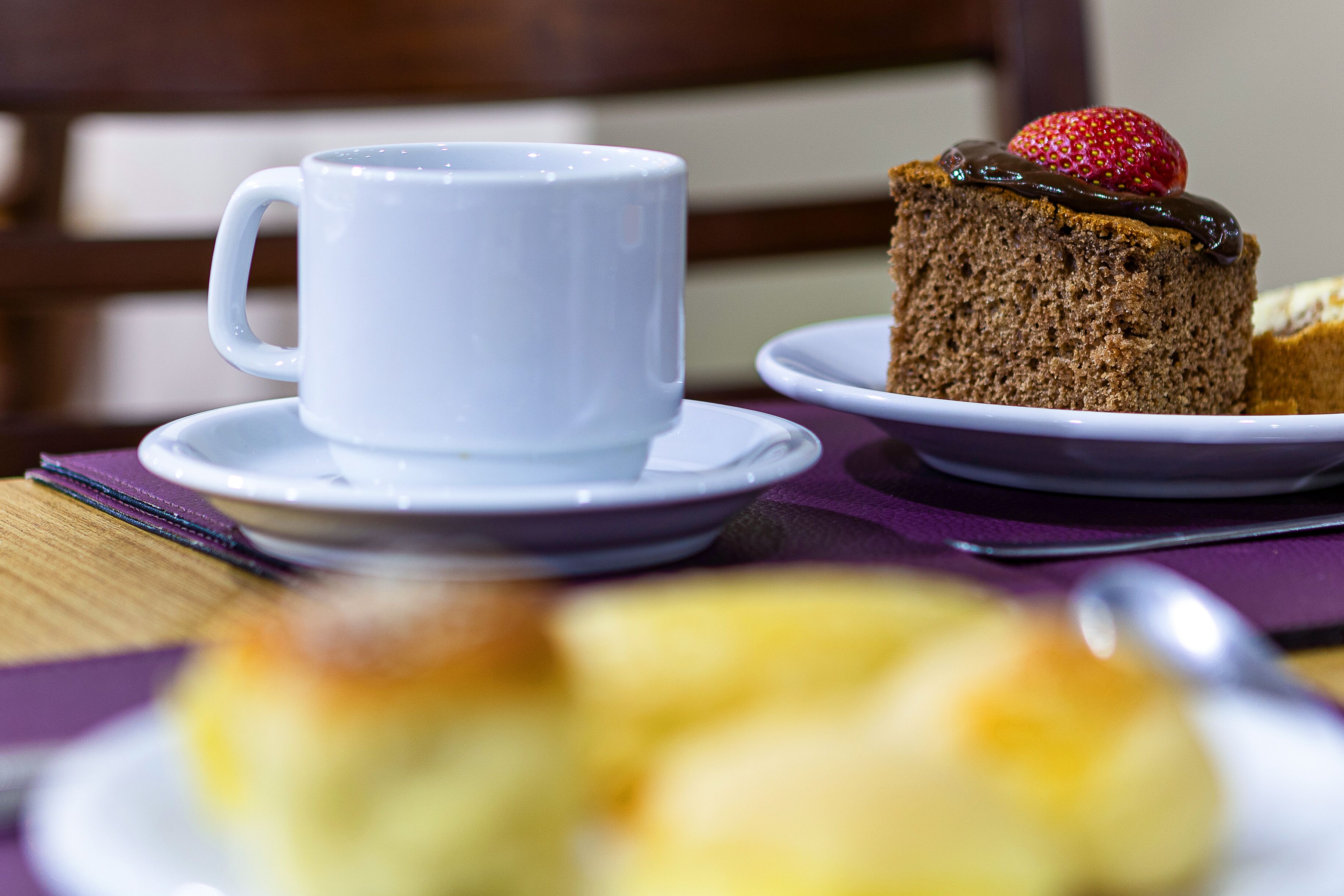 A white cup and saucer on a purple tablecloth, a slice of cake with a strawberry on top, and a blurred dessert in the foreground.