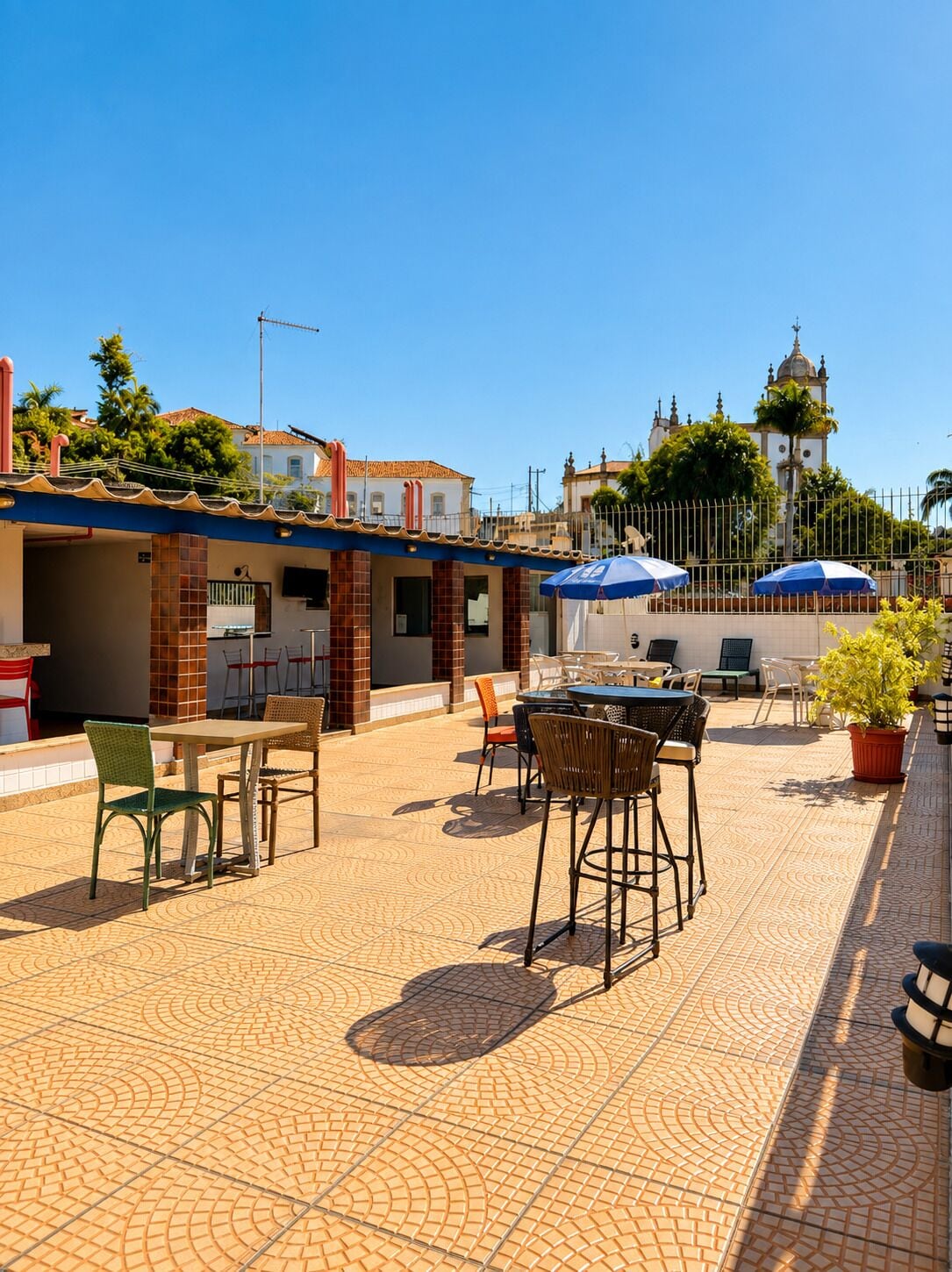 Un patio exterior con mesas y sillas, piso de baldosas y un edificio con un campanario al fondo.