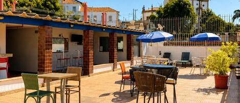 Un patio exterior con mesas y sillas, piso de baldosas y un edificio con un campanario al fondo.