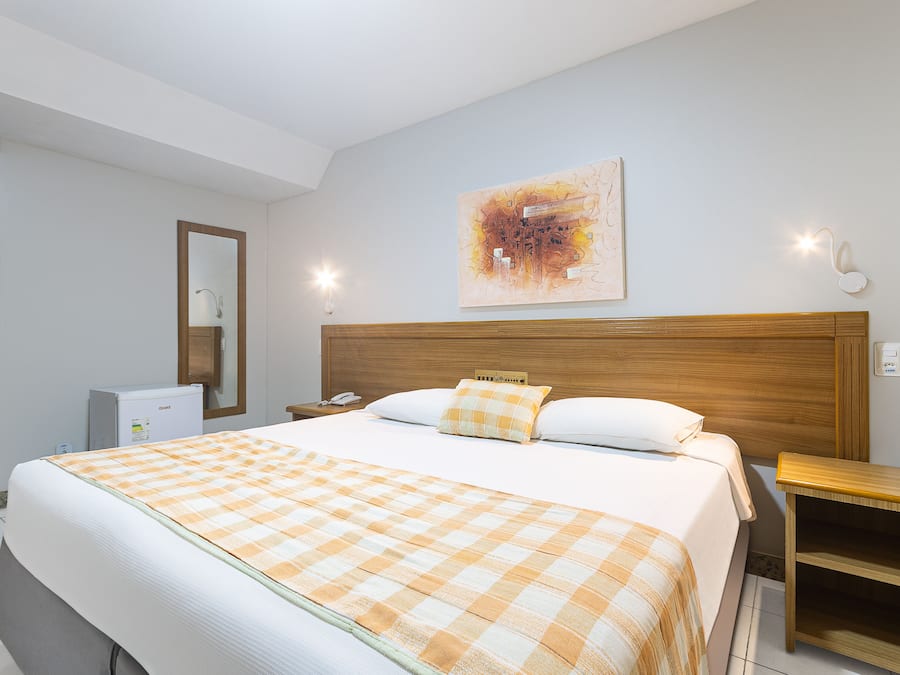 A hotel room with a large bed, a wooden headboard, a bedside table, a nightstand, a desk, a chair, a mirror, and a wall-mounted artwork.