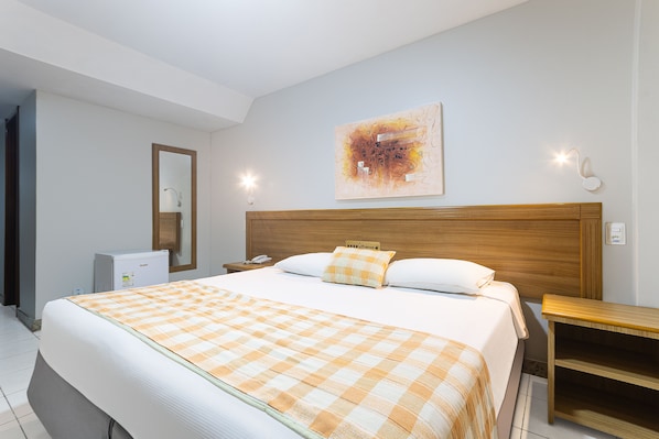 A hotel room with a large bed, a wooden headboard, a bedside table, a nightstand, a desk, a chair, a mirror, and a wall-mounted artwork.