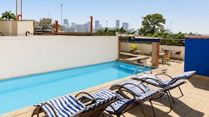 Une piscine sur le toit, agrémentée de chaises longues, avec une vue sur les toits de la ville en arrière-plan et un ciel dégagé.