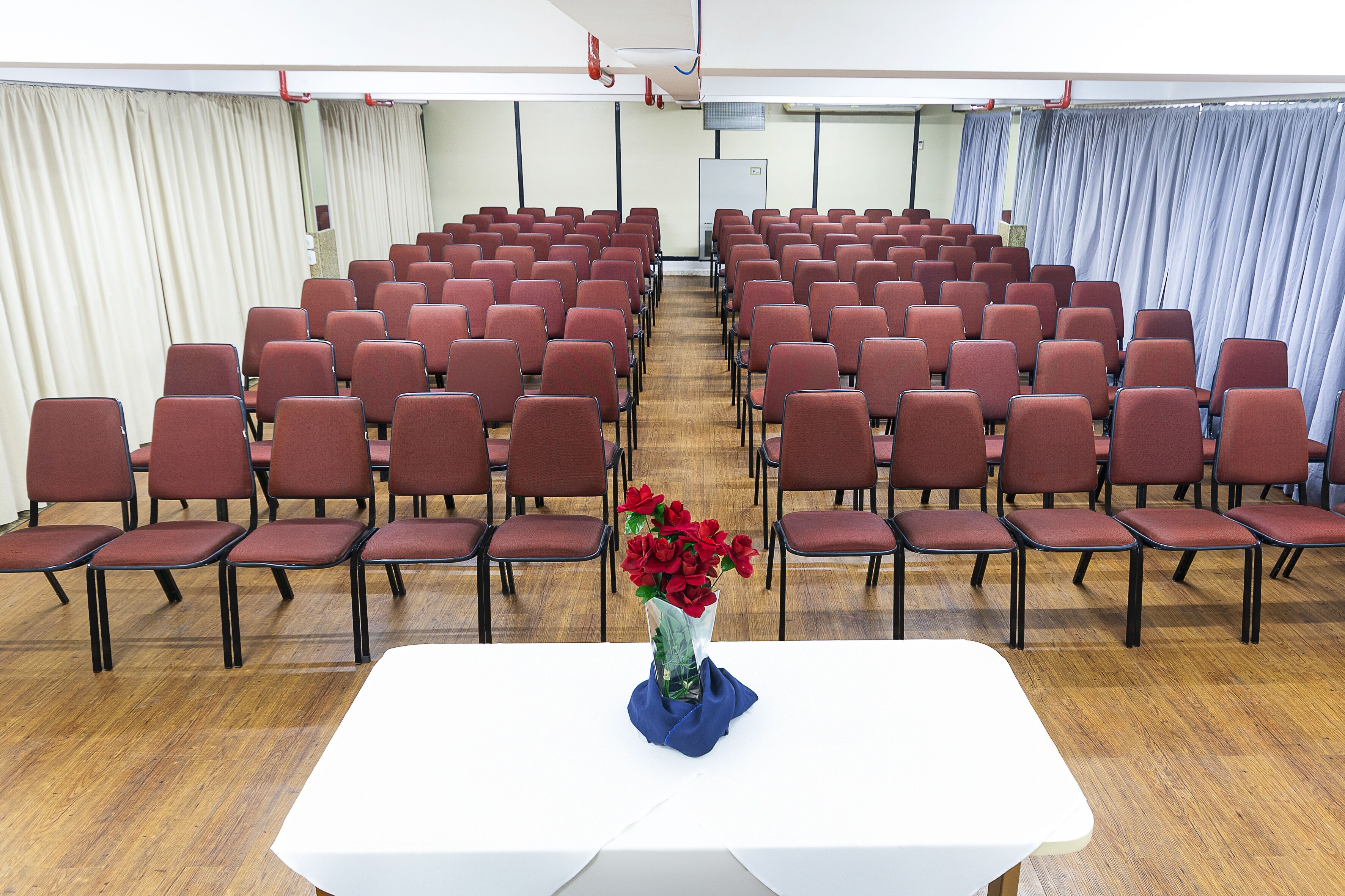 Une salle de conférence avec des rangées de chaises rouges et une table blanche ornée d’un vase rempli de fleurs rouges.