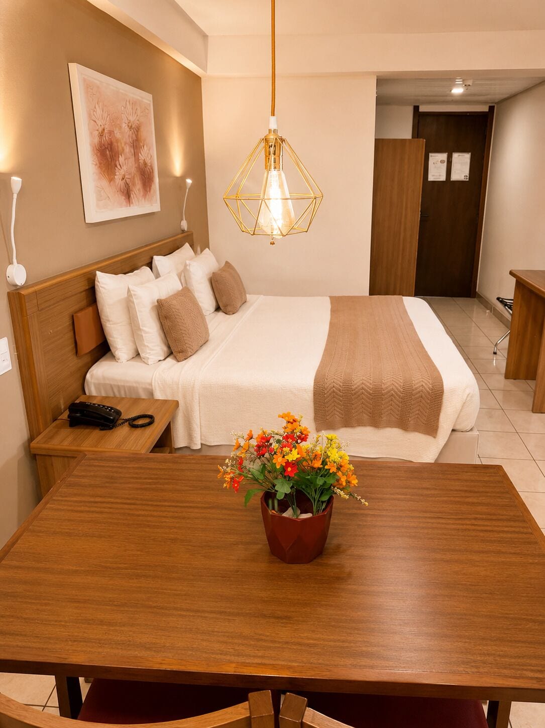 A hotel room with a bed, bedside table, lamp, and a dining table with a flower centerpiece.