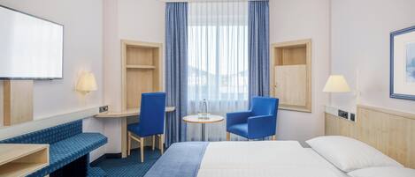 Habitación de hotel con una cama, dos sillas azules, una mesita redonda, televisión y una ventana con cortinas.