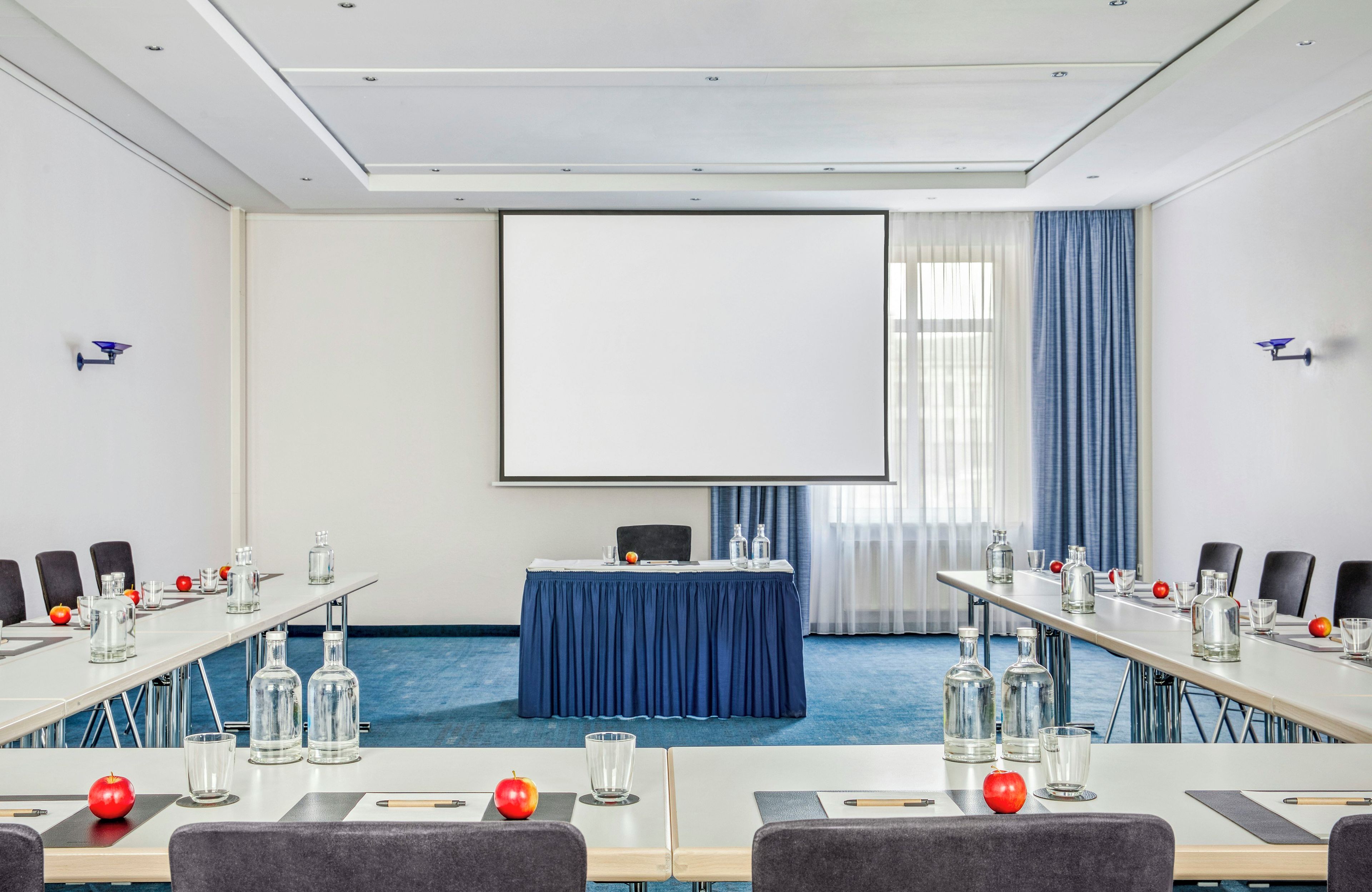 A conference room with a large screen, tables with chairs, and a blue carpet.