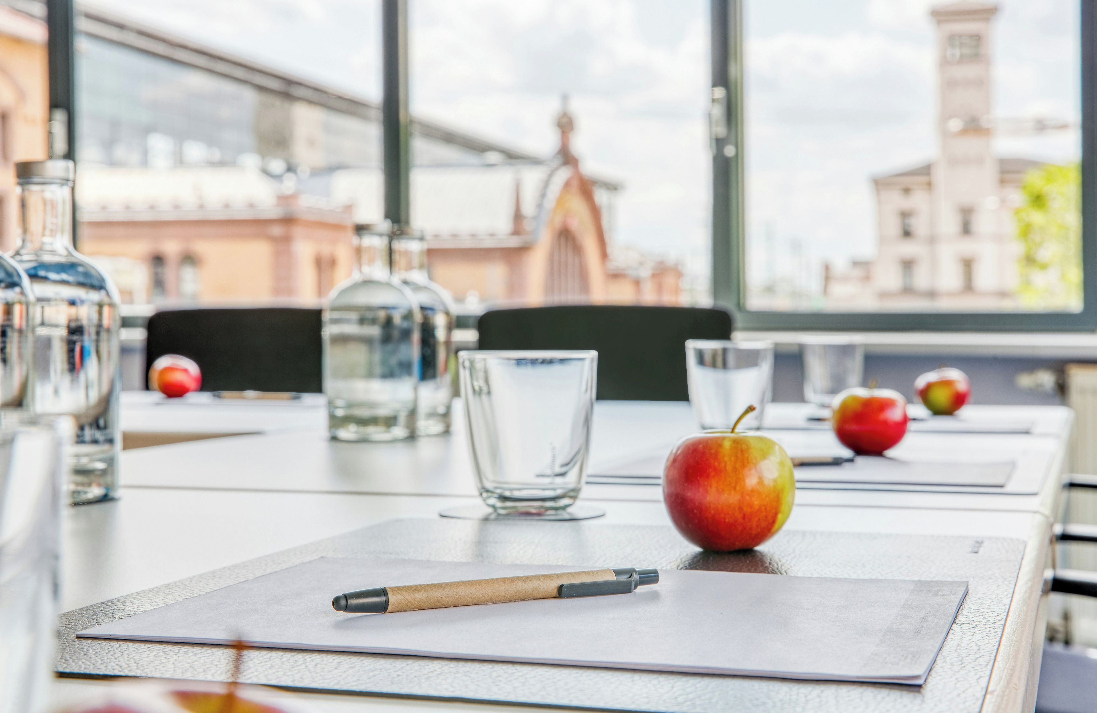 Ein Tisch mit Notizbuch, Stift, Brille und Äpfeln, durch große Fenster ist die Stadt zu sehen.