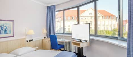A hotel room with a bed, desk, chair, and large windows offering a view of buildings.