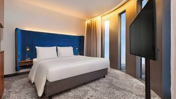 A hotel room with a large bed, a flat-screen TV, a nightstand, and a view of the city through large windows.