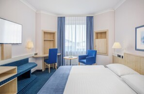 A hotel room with a large bed, two blue chairs, a small table, and a TV on the wall. - Hotel Erfurt City Center by Leonardo Hotels (Erfurt)
