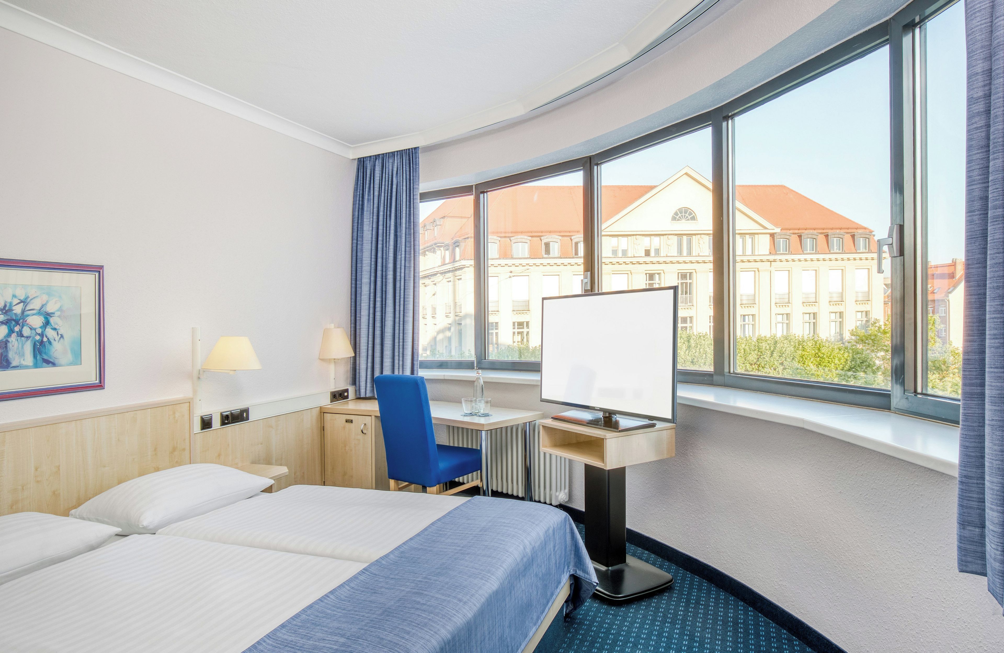 Una habitación de hotel con cama, escritorio, silla y un amplio ventanal con vistas a edificios.