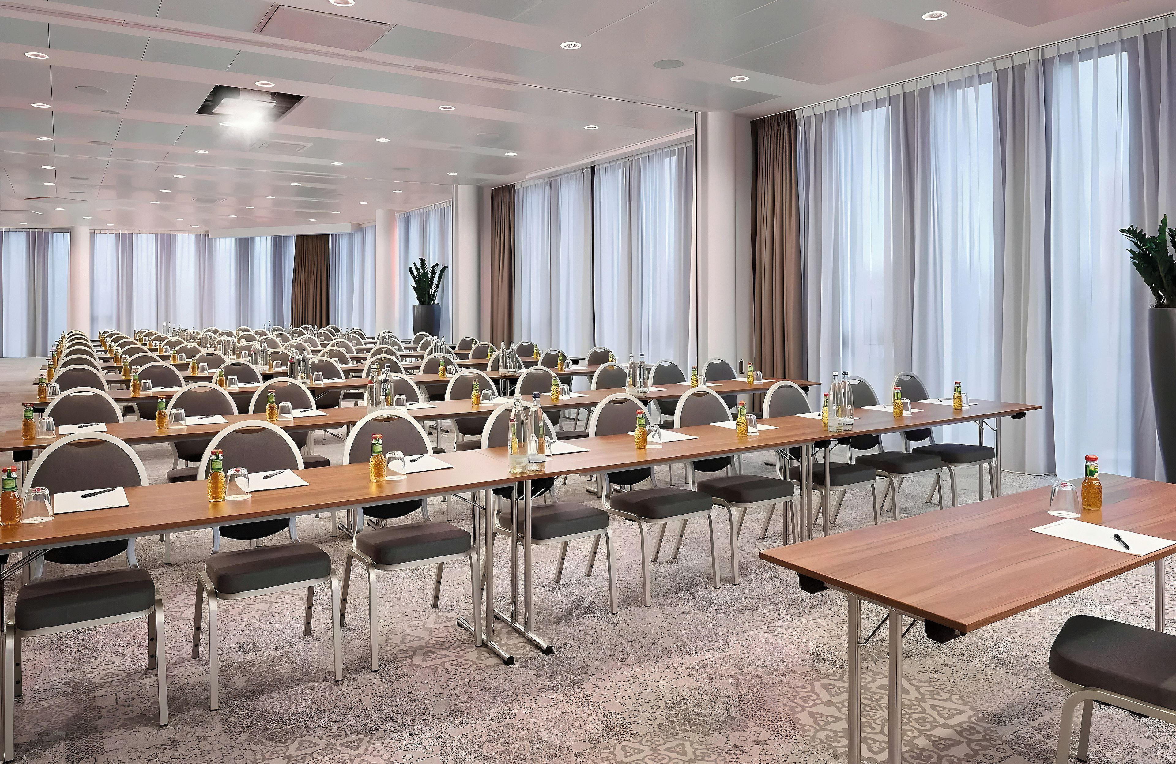 Une salle de conférence dotée de plusieurs tables et de chaises, de grandes fenêtres avec des rideaux et d’un tapis à motifs.