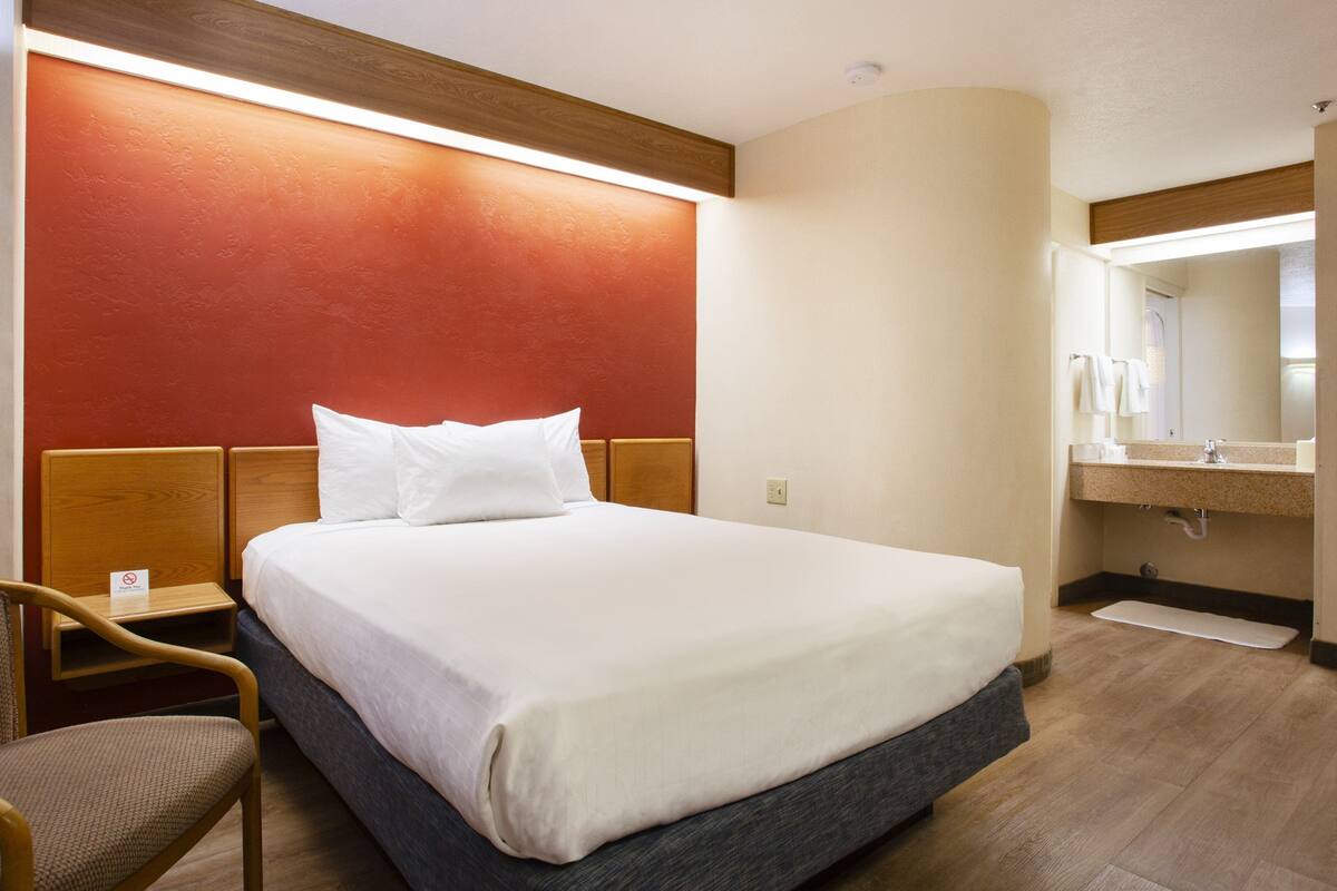 A hotel room with a bed, a chair, a nightstand, a bathroom with a sink and mirror.