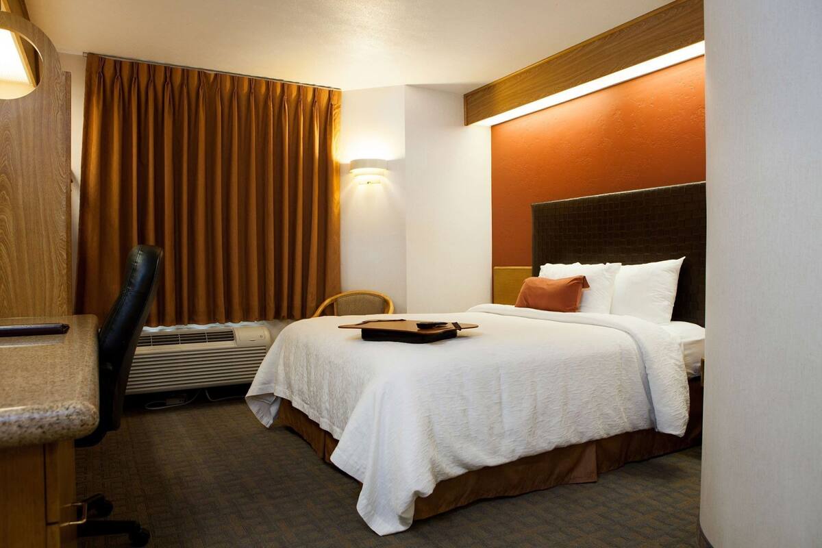 A hotel room with a bed, a desk, a chair, a lamp, and a wall-mounted heater.