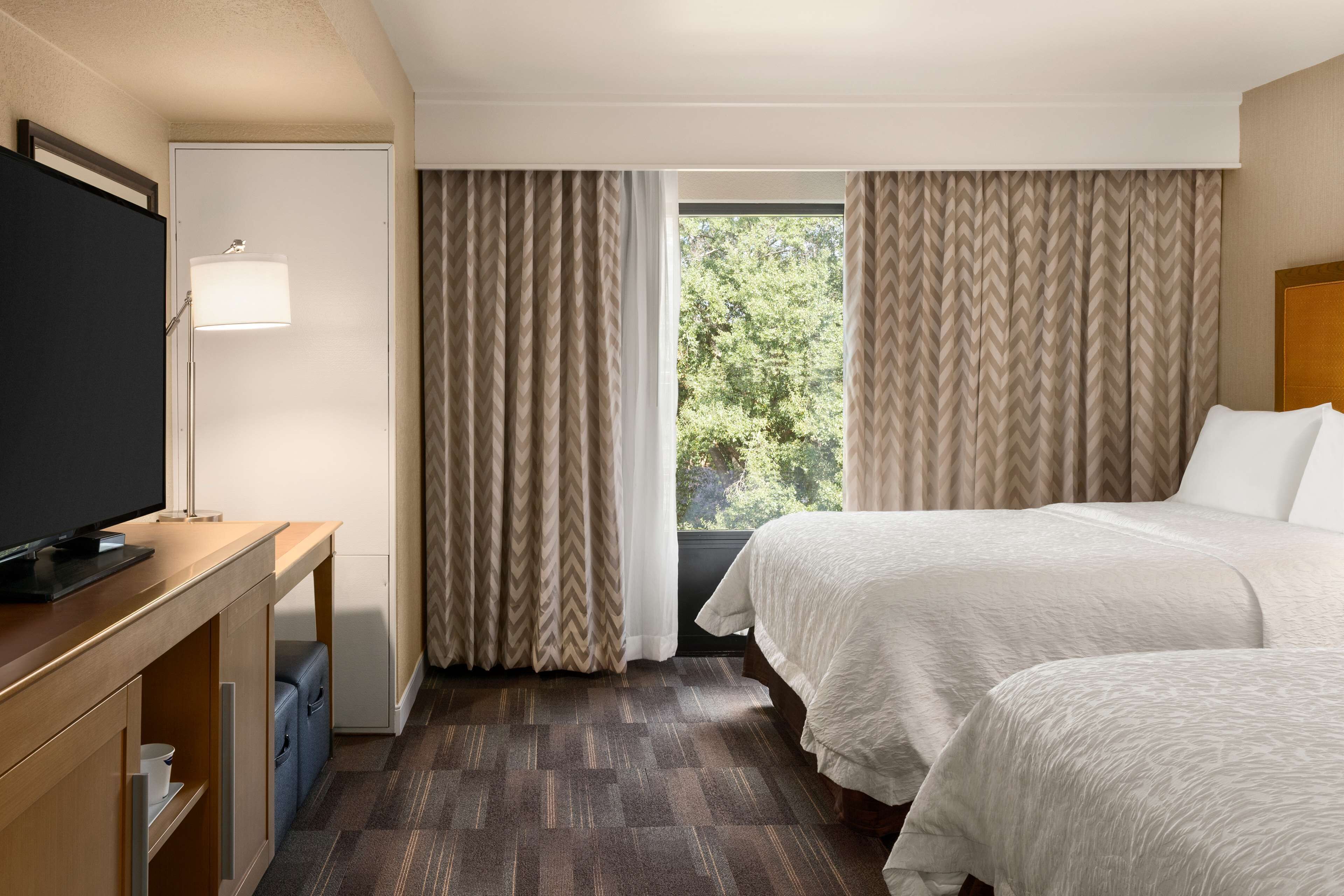 Ein Hotelzimmer mit zwei Betten, einem Schreibtisch und einem Fernseher sowie einem Fenster mit Vorhängen.