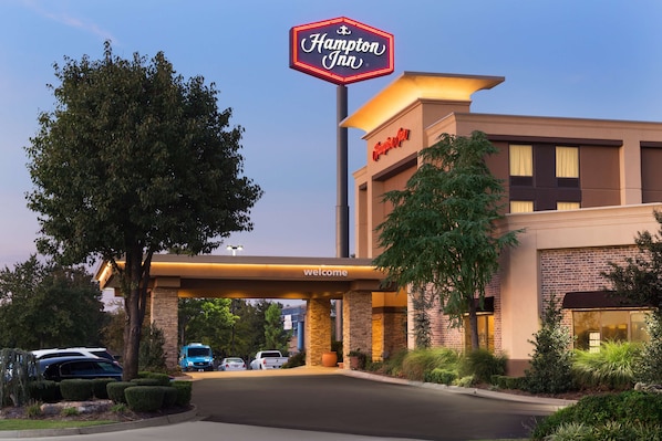 A Hampton Inn hotel entrance with a 'Welcome' sign, a parking lot, and a clear sky.