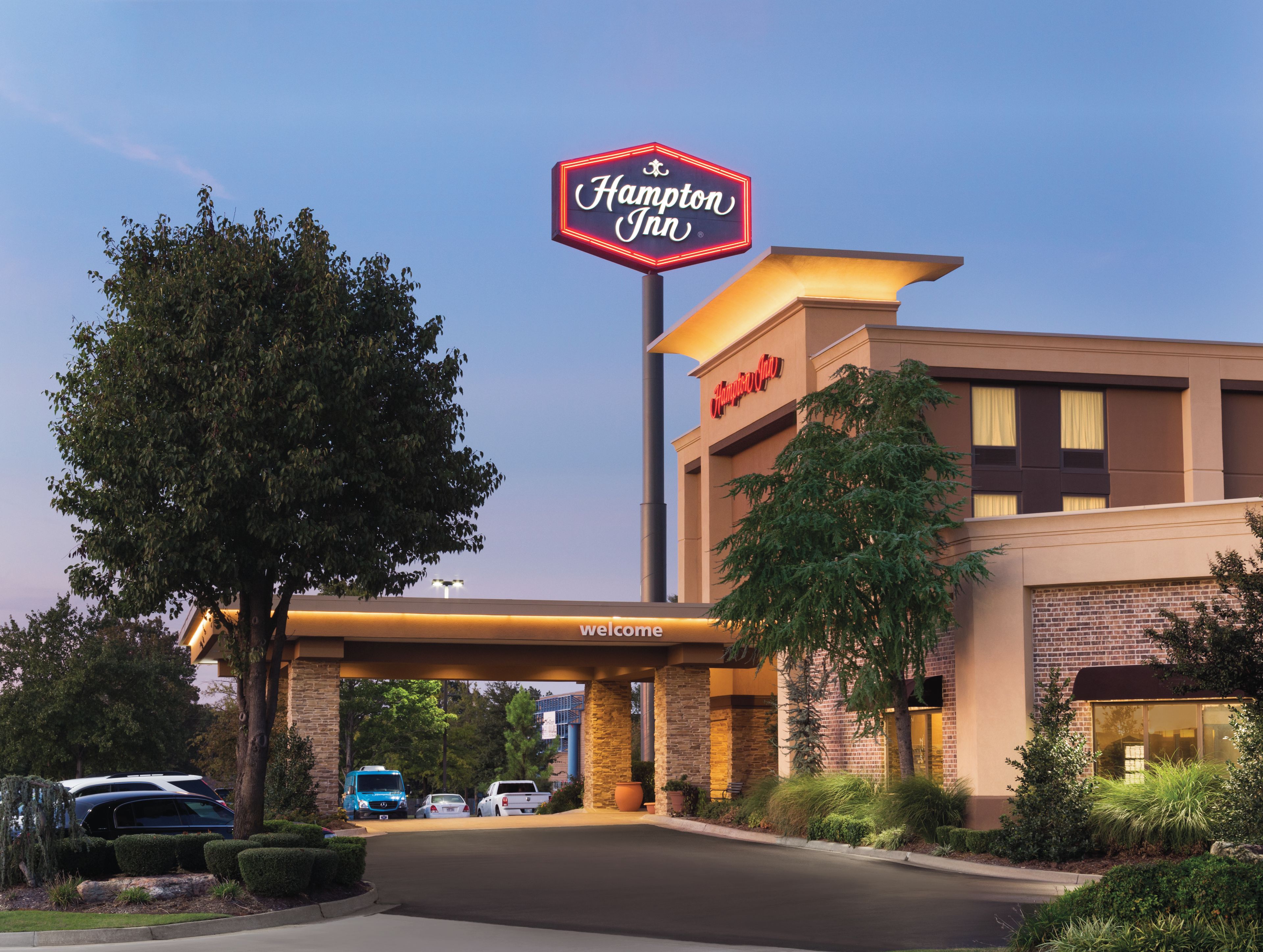 A Hampton Inn hotel with a clear sky, a welcoming sign, and a parking lot with several vehicles.