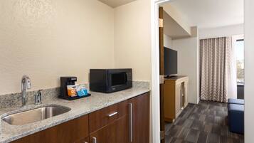 A compact kitchen with a granite countertop, stainless steel sink, and a microwave. There is a small coffee maker and a television on a stand. The room has wooden cabinets and a carpeted floor.