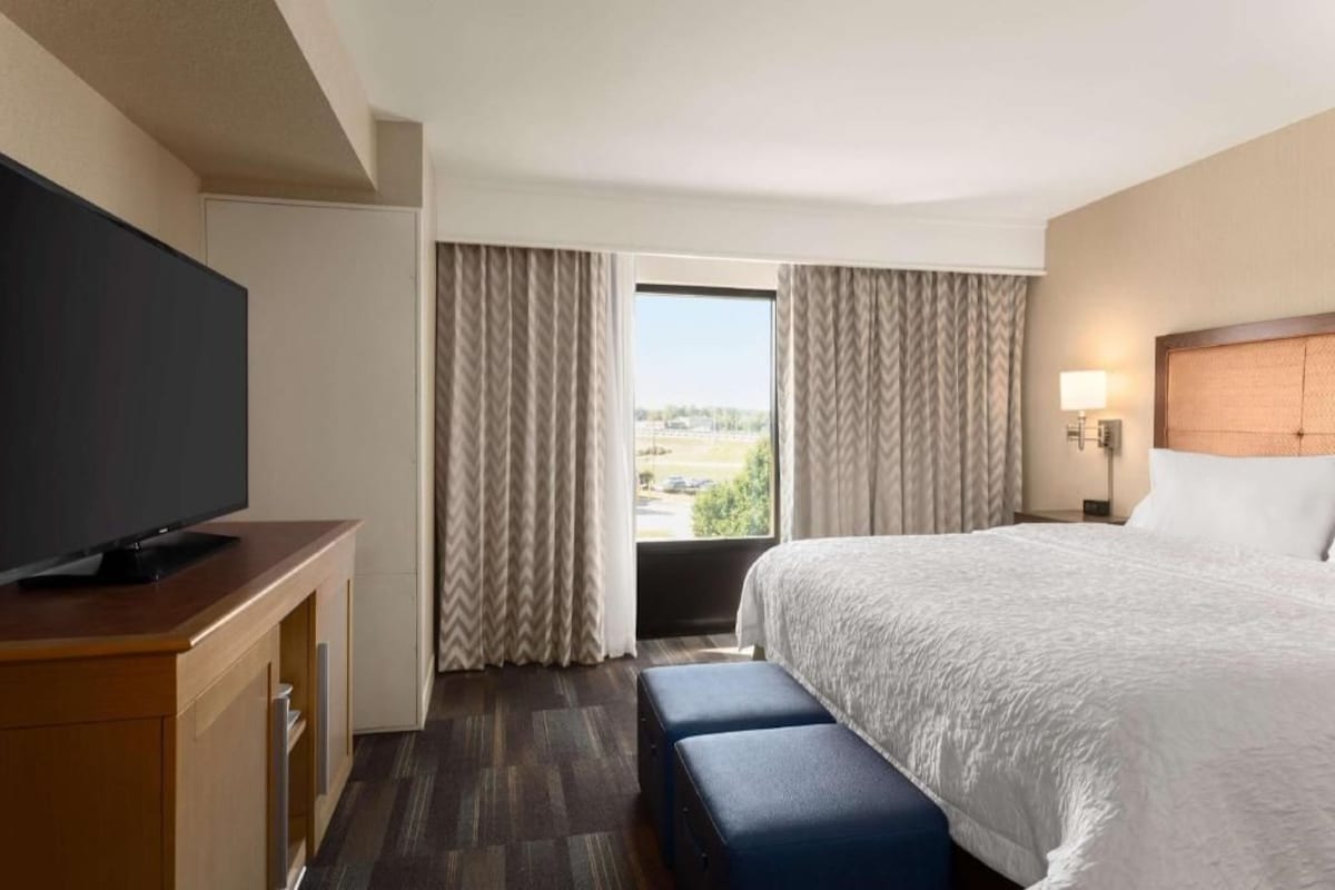 Habitación de hotel con cama, televisor sobre un mueble de madera, ventana con cortinas y lámpara en la mesita de noche.