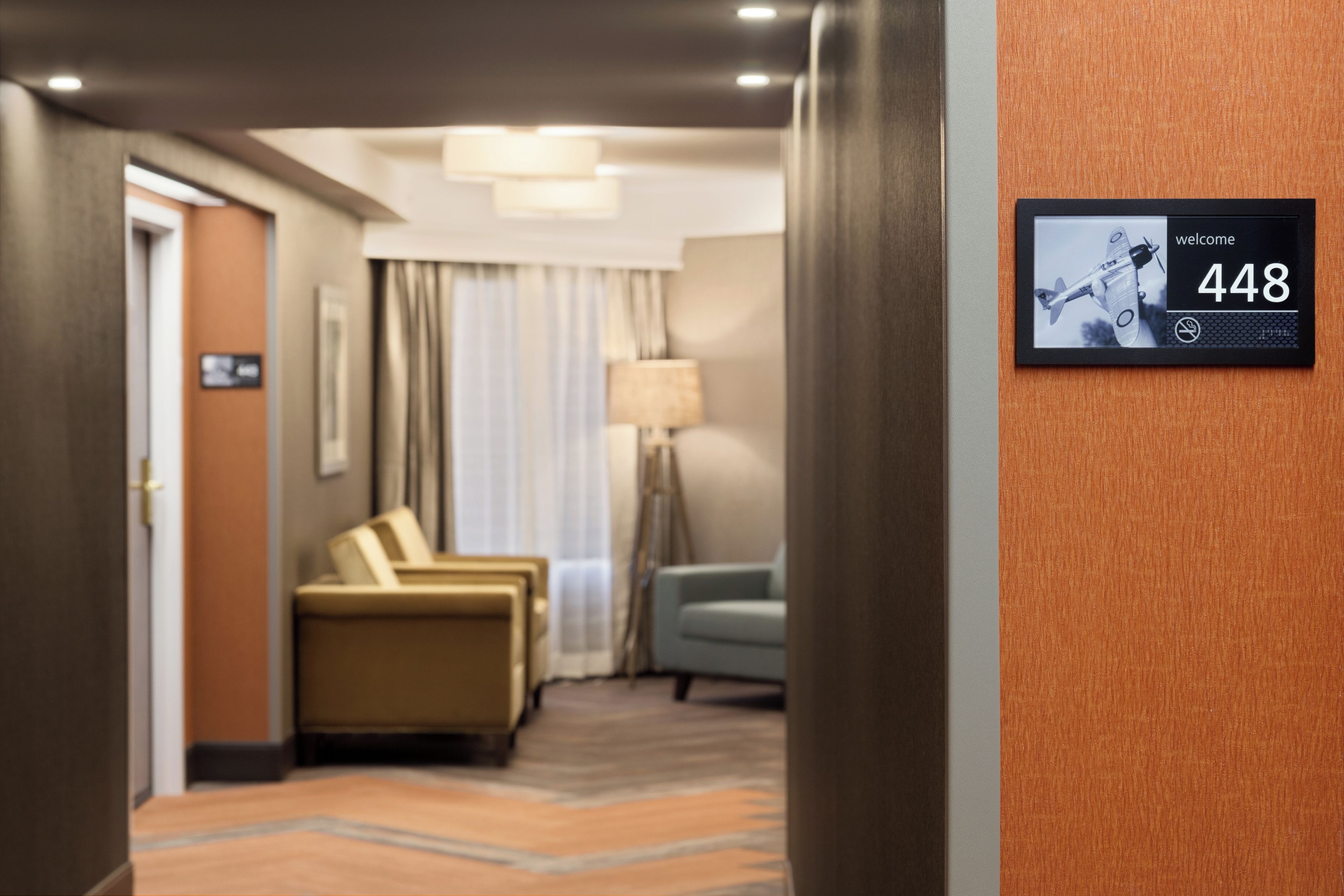Un couloir d’hôtel avec un comptoir de réception, un espace détente avec un canapé et des fauteuils, et un écran numérique fixé au mur affichant le numéro de chambre 448.