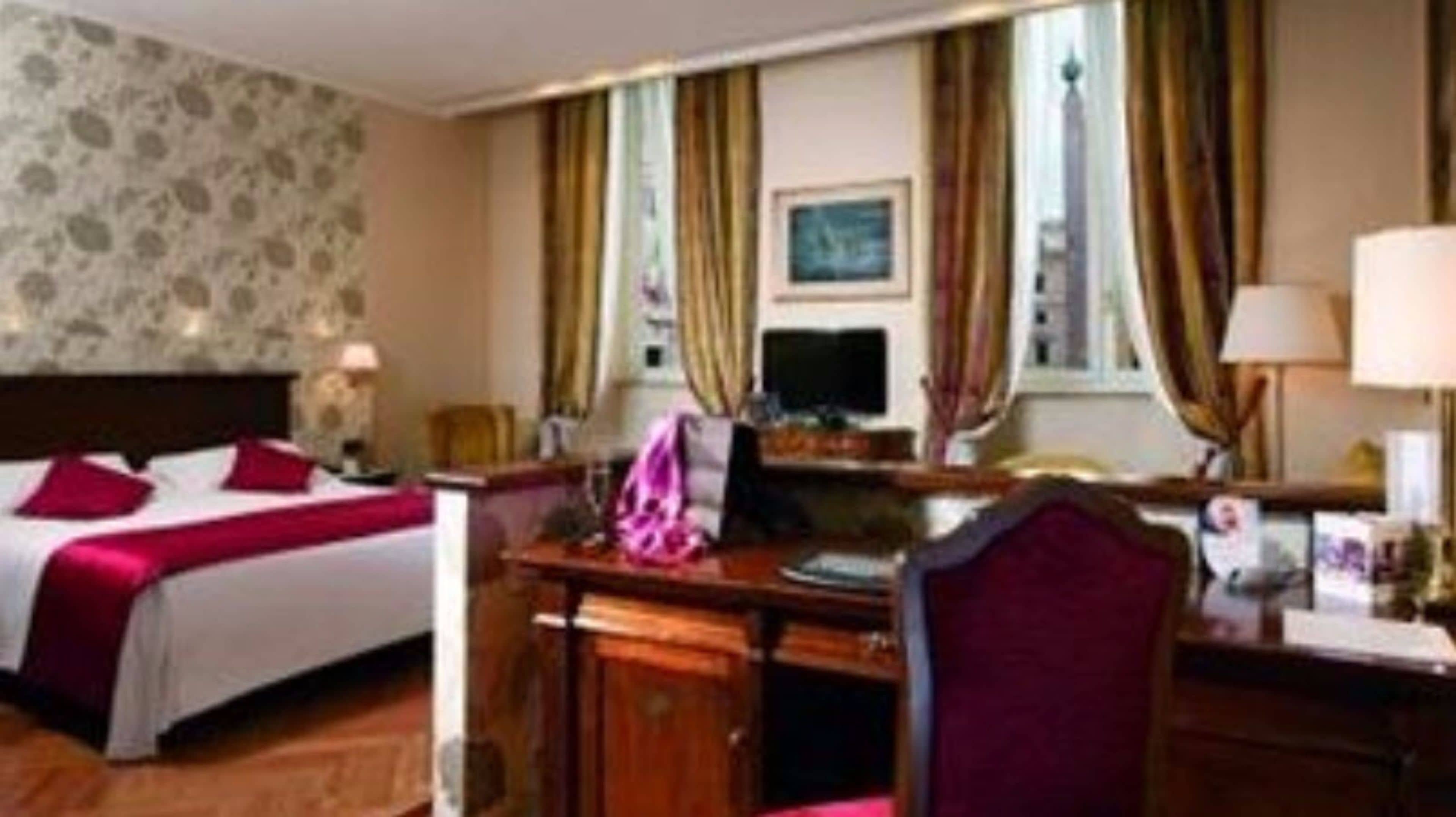 Quarto de hotel com cama, escrivaninha e cadeira. Tem uma janela com cortinas, uma luminária e um quadro na parede.