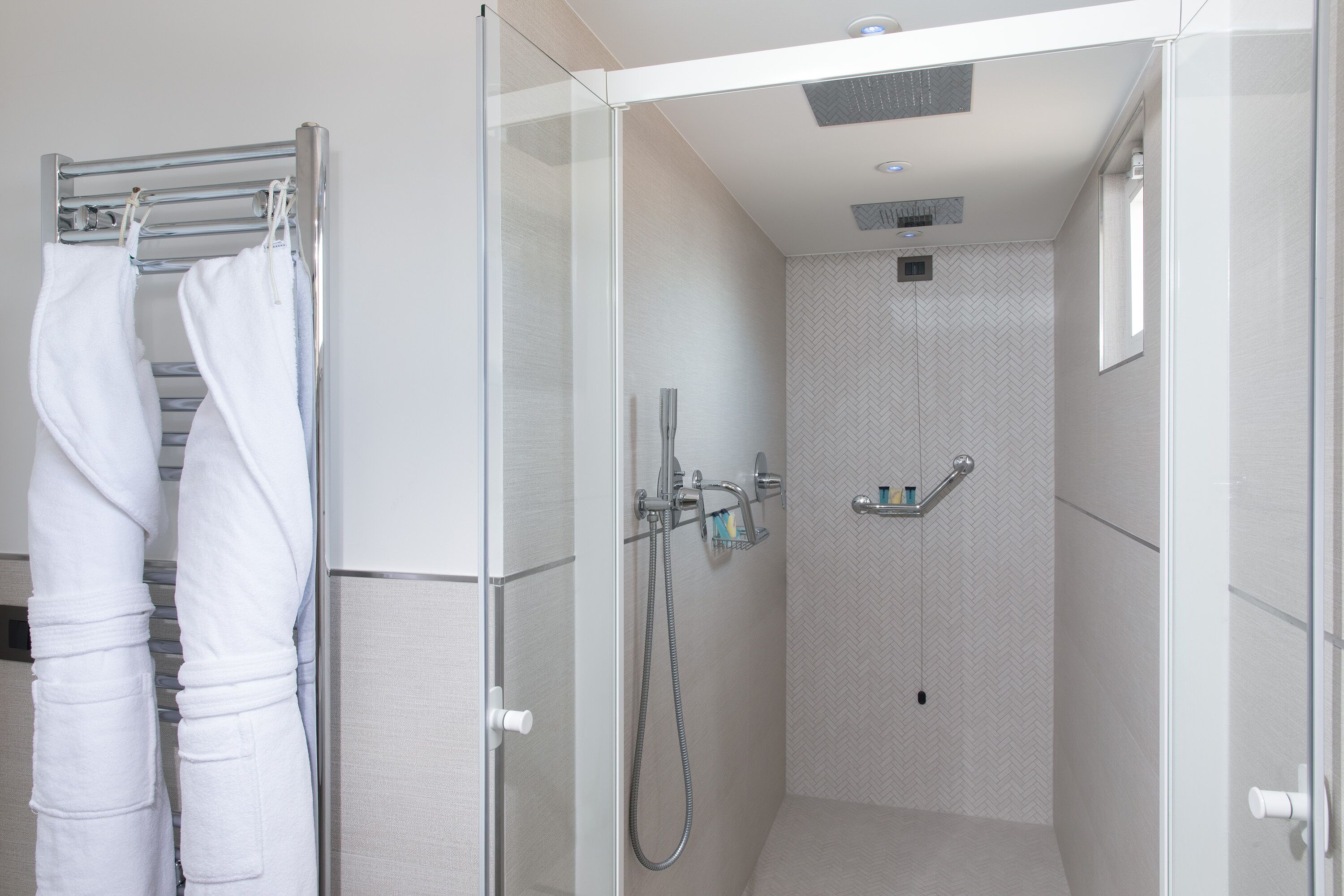 A modern shower area with a glass door, a towel rack with white towels, a handheld showerhead, and a handheld soap dispenser.