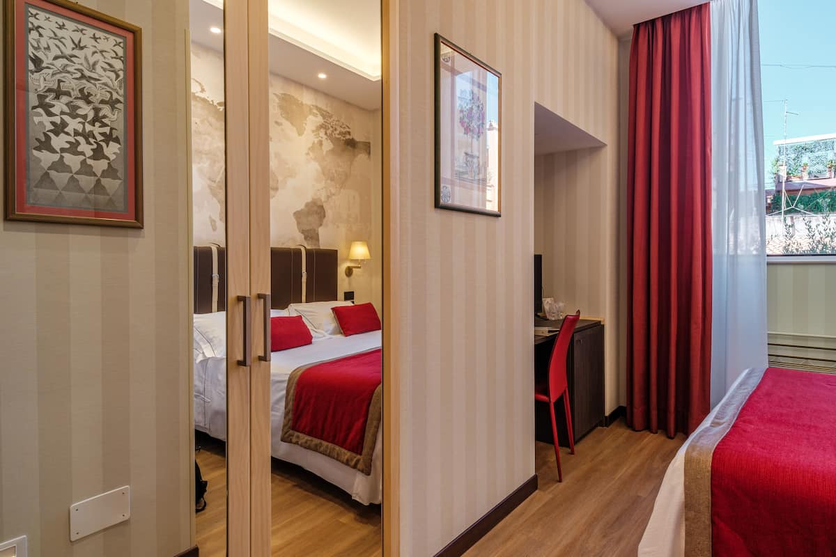 Ett hotellrum med en säng, ett skrivbord och en stol, ett stort fönster med röda gardiner och konstverk på väggarna.