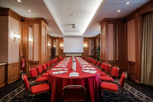 Een vergaderzaal met een lange tafel, rode stoelen en een projectiescherm aan de voorkant.