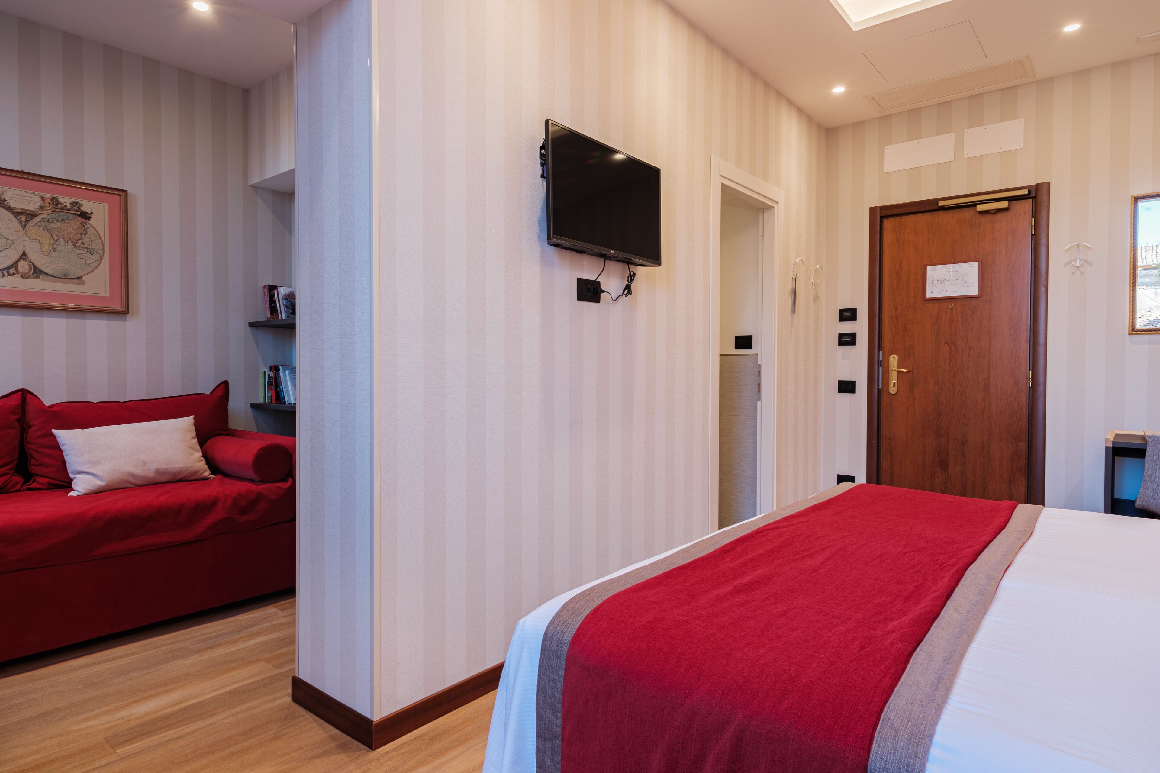 A hotel room with a bed, a red sofa, a TV mounted on the wall, and a wooden door.
