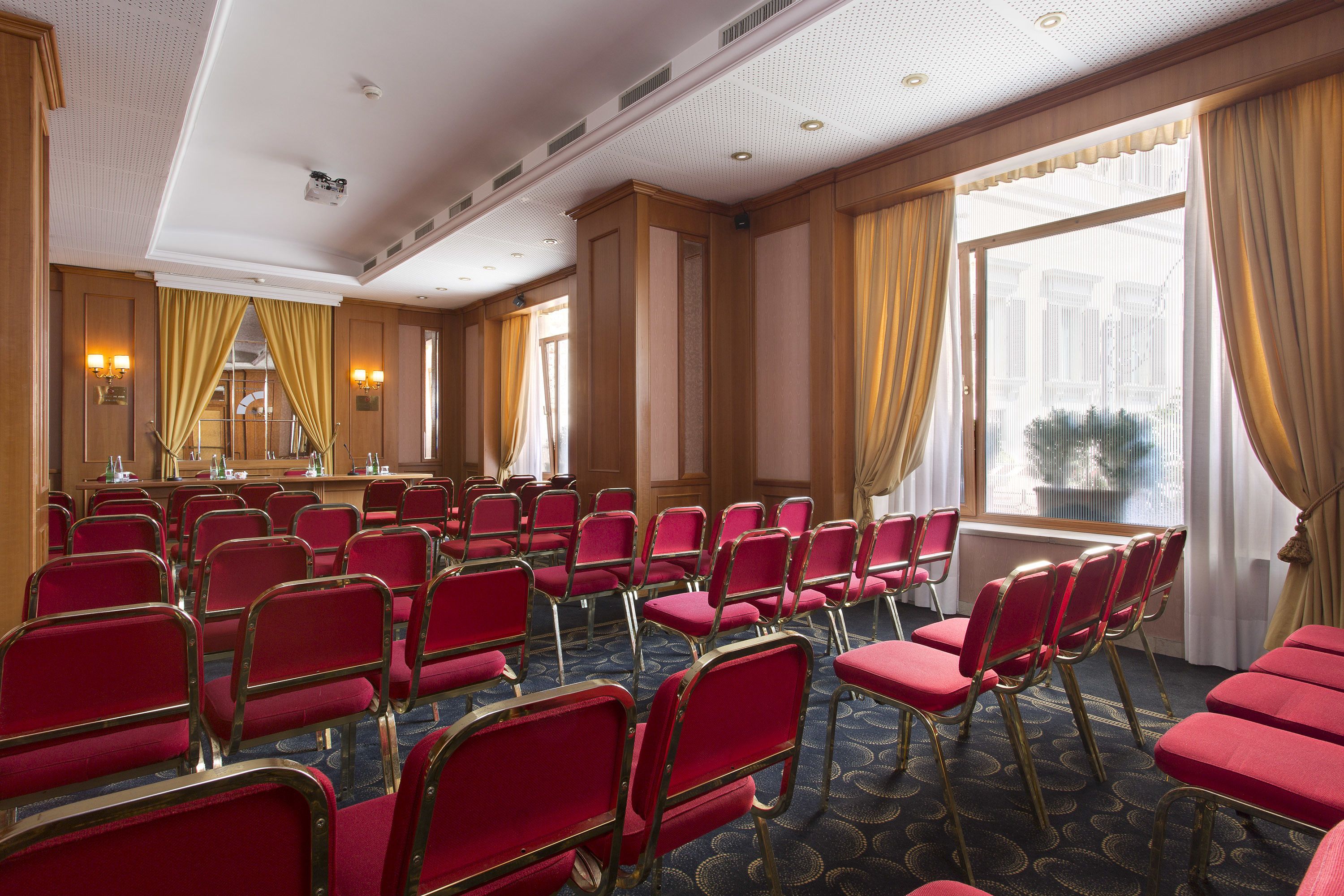 A conference room with rows of red chairs, a podium, and large windows with curtains.
