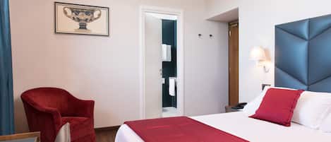 Habitación de hotel con una cama, un sillón rojo, un cuadro en la pared y una puerta que da a un baño.