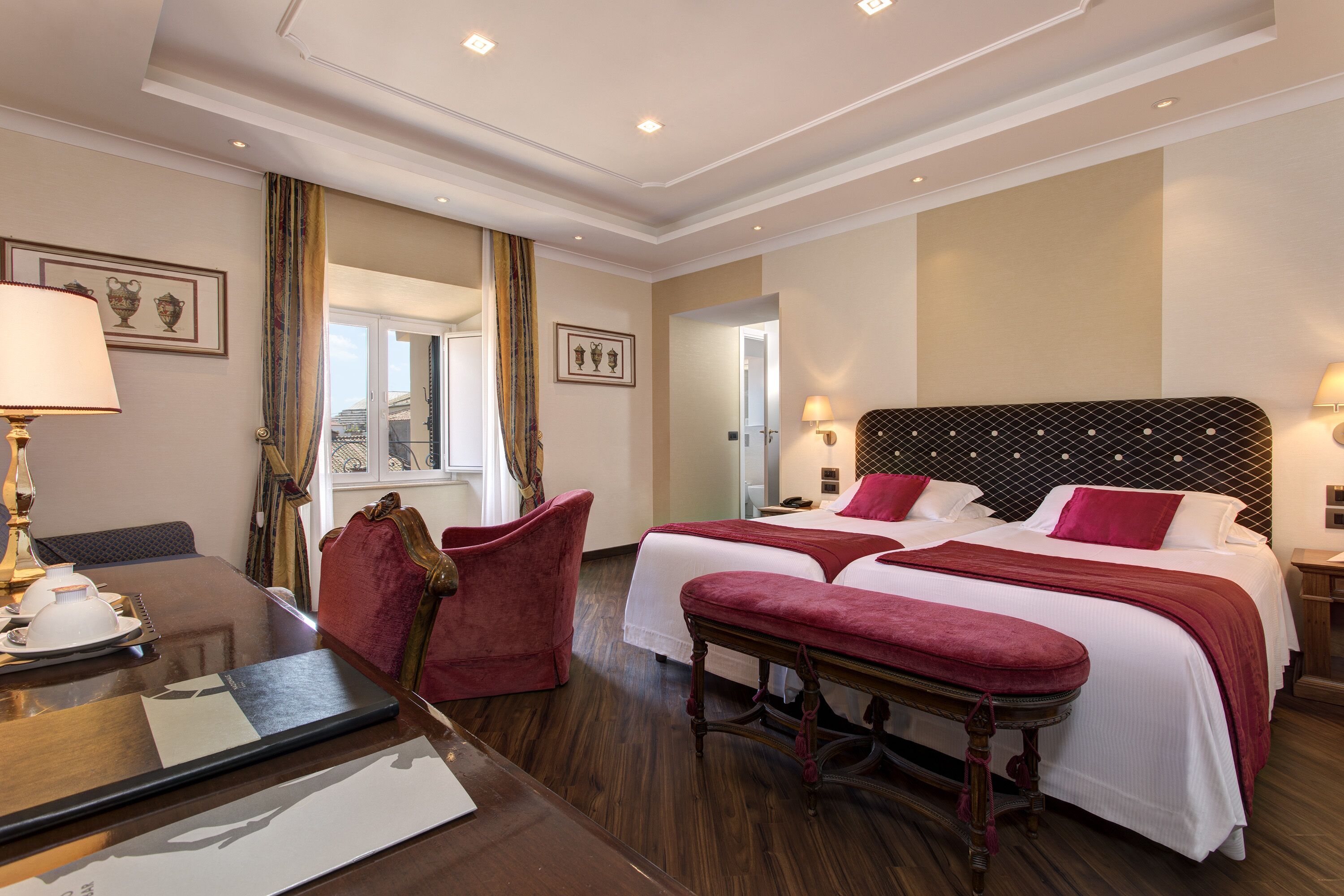 Une chambre d’hôtel avec un plancher en bois, deux lits avec une literie rouge, un bureau avec une lampe et une chaise.