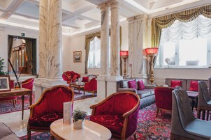 A lobby with marble columns, red upholstered chairs, a patterned carpet, and a red lamp.