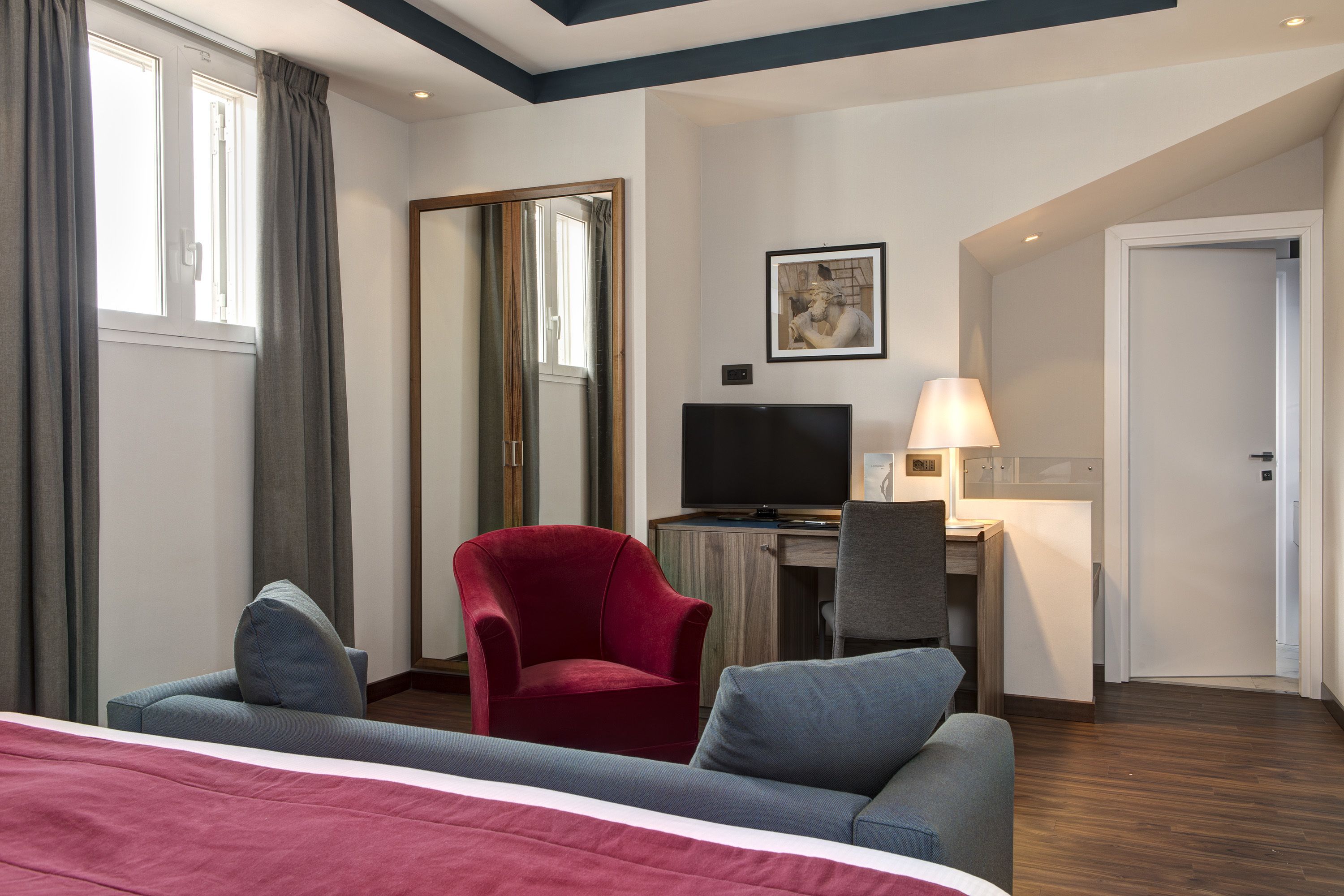 A hotel room with a bed, a desk with a computer, a red armchair, a grey armchair, a TV, and a door.