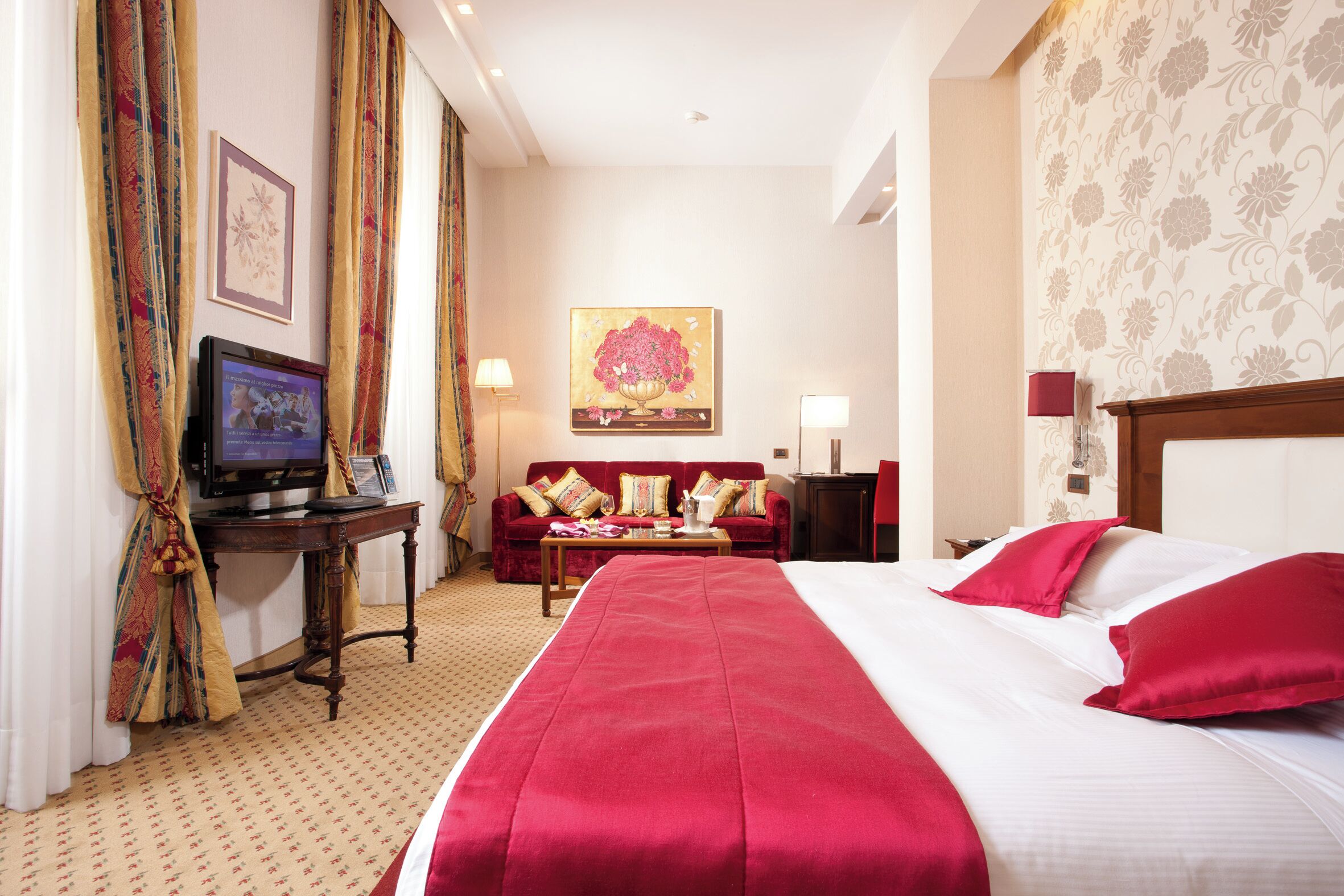 A hotel room with a large bed, a desk with a computer, a red sofa, and a painting on the wall.
