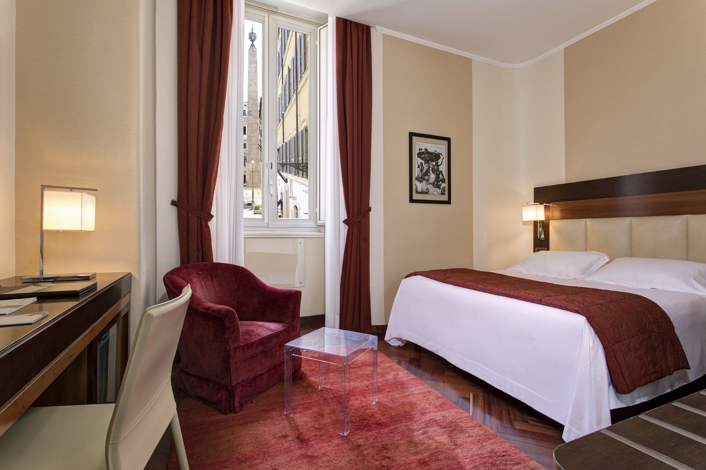 Une chambre d’hôtel comprenant un lit, un bureau avec une chaise, une petite table et une fenêtre avec des rideaux.