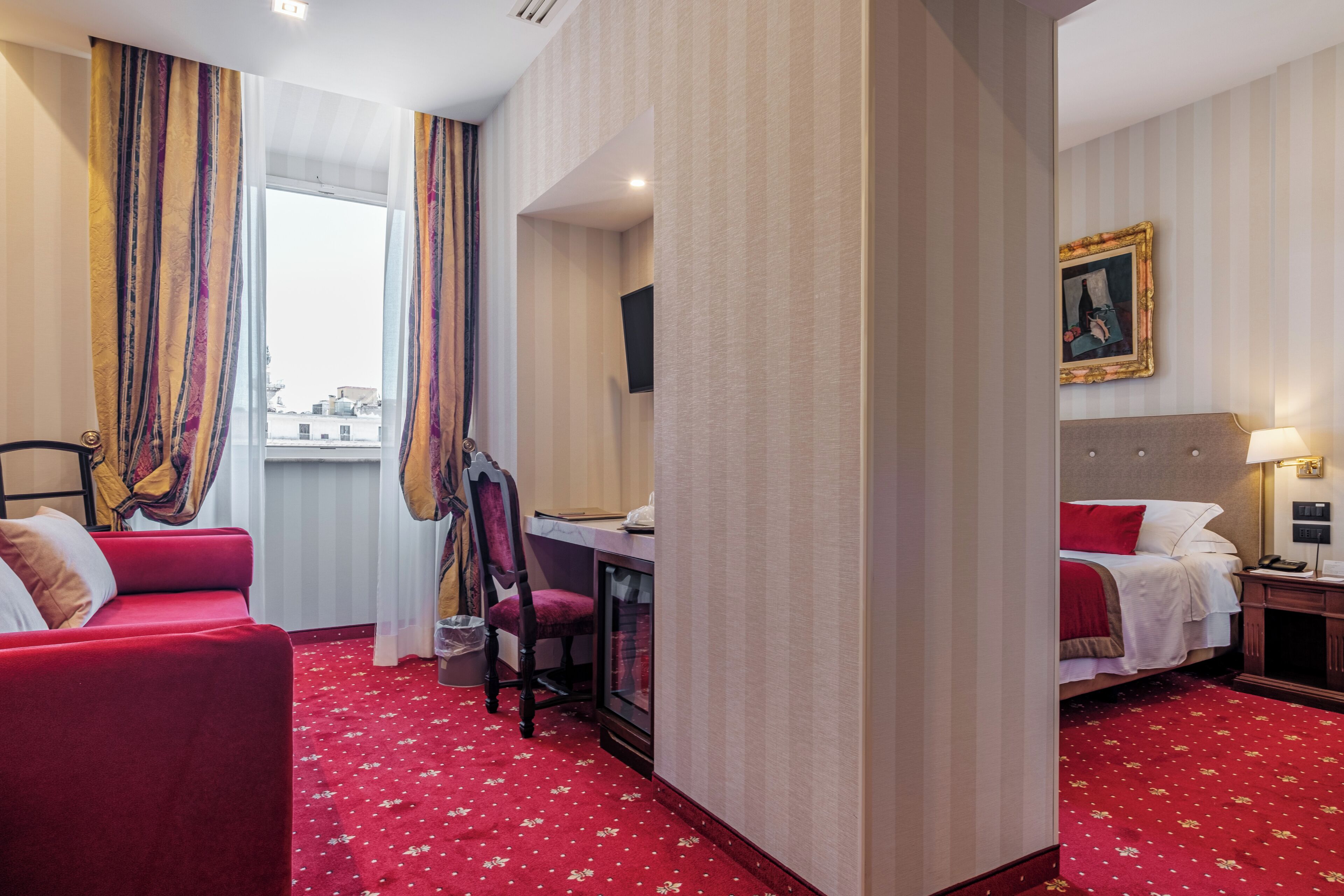 Een hotelkamer met een rode loper, een bureau met stoel, een bed met een rood kussen en een schilderij aan de muur.