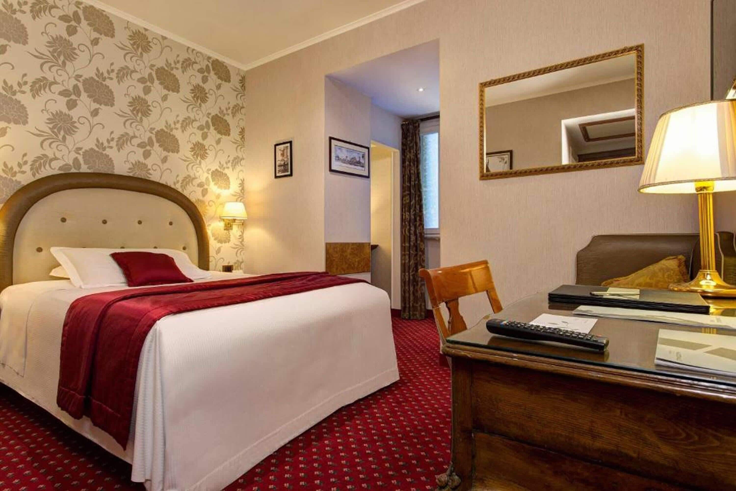 A hotel room with a large bed, a desk with a lamp, a chair, and a mirror on the wall.