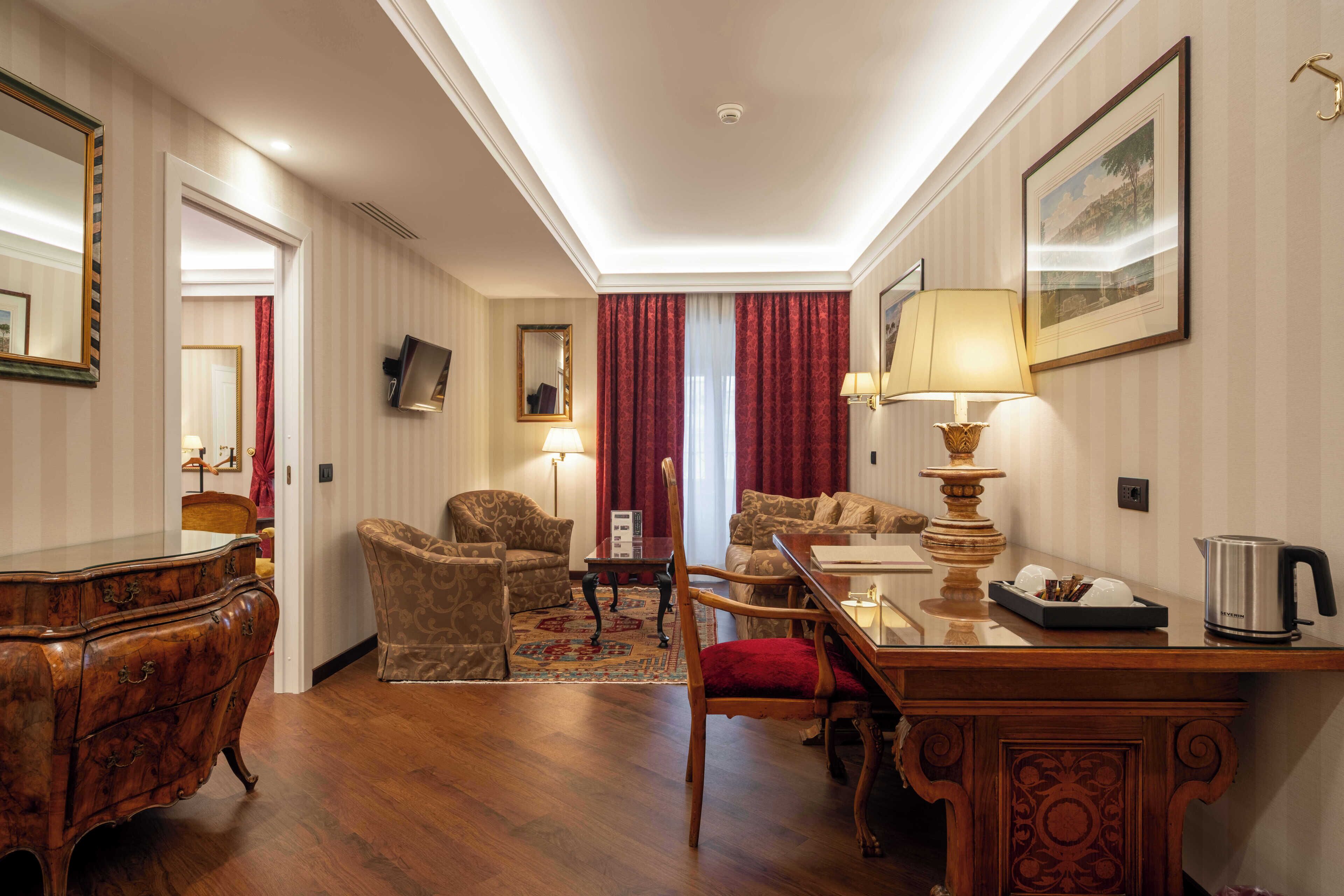 A hotel room with a wooden dining table, two chairs, a sofa, a red chair, a wooden dresser, and a television on the wall.