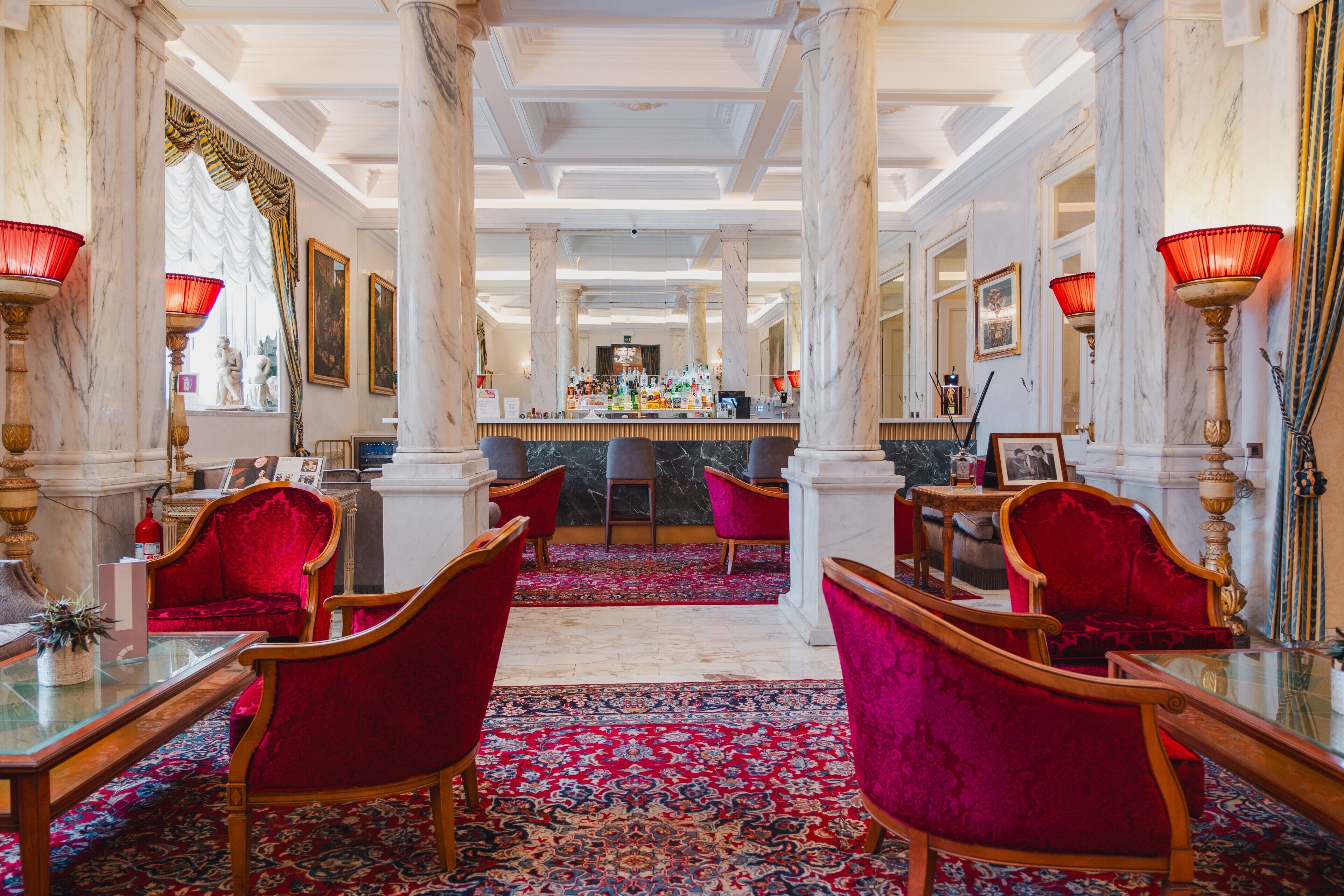 A hotel lobby with marble columns, red velvet chairs, a bar area, and a patterned carpet.