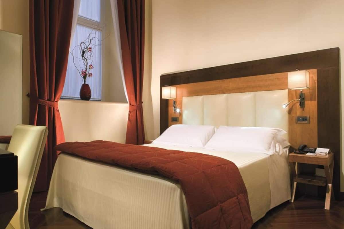 Ett hotellrum med en säng, ett nattduksbord, en stol, ett fönster med gardiner och en vas med blommor.
