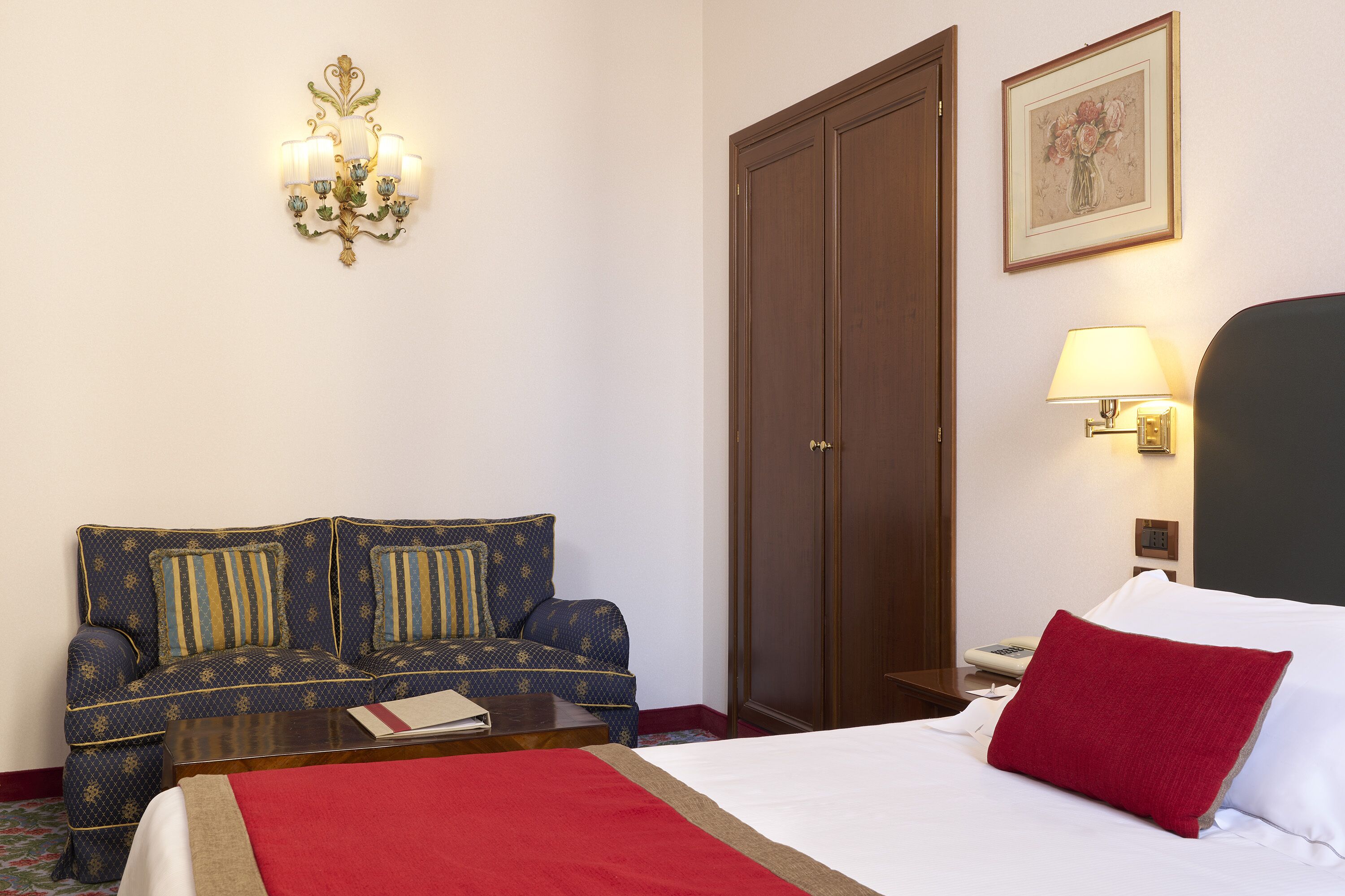 A hotel room with a bed, a sofa, a wooden wardrobe, a wall-mounted lamp, and a framed picture.